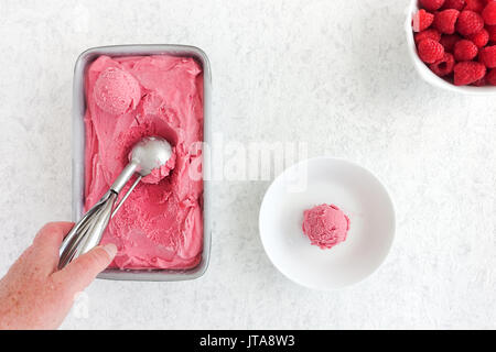 Mano fuori di movimentazione in casa gelato al lampone da una vasca metallica. Cucchiaio di gelato in una ciotola e lamponi freschi sul marmo bianco dello sfondo. Foto Stock
