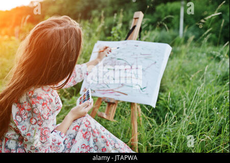 Ritratto di una splendida felice giovane donna nel bel vestito seduto sull'erba e pittura su carta con acquerelli. Foto Stock