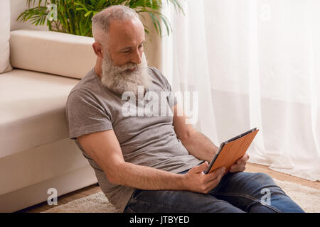 Senior uomo barbuto e sorridente con tavoletta digitale e seduto sul pavimento a casa Foto Stock