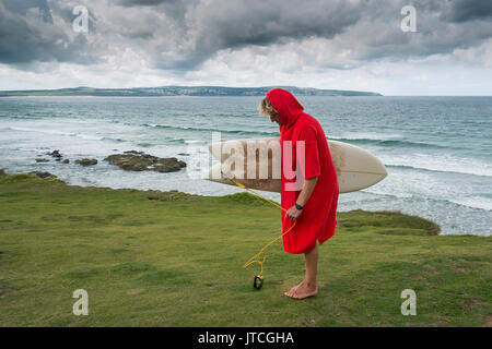 Un surfista indossando un rosso surf poncho tenendo una tavola da surf a Godrevy in Cornovaglia. Foto Stock