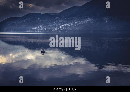 Idilliaco paesaggio di montagna con uomo in barca da pesca nel mezzo di un tranquillo lago con acqua riflessioni durante il sunrise con drammatico nel cielo blu a Foto Stock