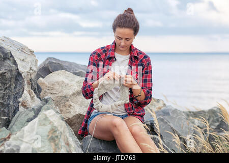 Una giovane ragazza dai capelli scuri in jeans e una a scacchi in maglietta rossa a maglia un maglione grigio dal naturale vestiti di lana e rocce sulle pietre vicino al mare befo Foto Stock