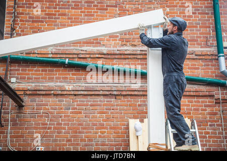 Un giovane uomo con la barba builder in una costruzione di colore nero uniforme, un cappuccio grigio costruisce una casa di una trave di legno dipinto di bianco in un giorno di estate, la BAC Foto Stock