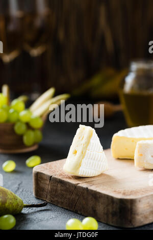 Francese di formaggio morbido dalla regione della Normandia affettata con verde uva, pera, miele e bicchieri di vino su una tavola di legno scuro su sfondo rustico Foto Stock