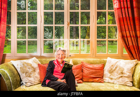 Donna anziana sorridente che si rilassa nel suo salotto tenendo un libro con grande finestra al giardino dietro Foto Stock