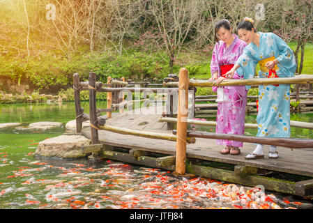 Bella donna giapponese medicazione kimono al sakura parco giardino godendo di guardare molto pesce carpe nella parte anteriore del laghetto di acqua sul percorso di legno e puntamento c Foto Stock