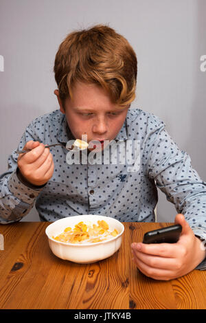 11-anno vecchio ragazzo guardando i social media su un telefono intelligente mentre si consuma la prima colazione, Bawdsey, Suffolk, Inghilterra. Foto Stock