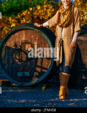 Primo piano su sorridente brunette donna enologo in piedi vicino a barile di legno in vigneto all'aperto in autunno. Small business concept Foto Stock