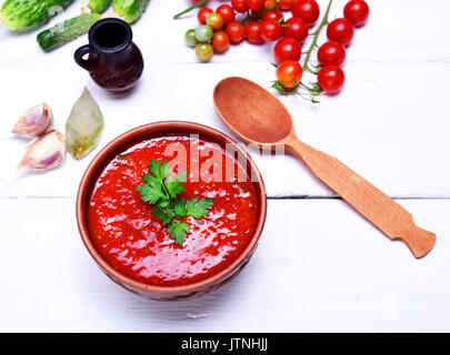 Zuppa Gazpacho in una piastra marrone su un bianco sullo sfondo di legno con un cucchiaio di legno, vista dall'alto Foto Stock