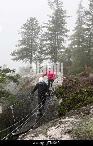 Un uomo e una donna si attraversa un piccolo ponte di sospensione che è parte della Via Ferrata in Squamish, British Columbia. Foto Stock