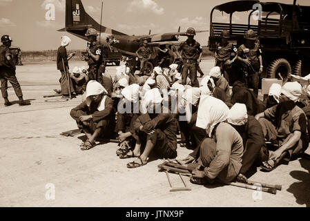 Il Viet Cong POWs sedersi sulla rampa a Tan Son Nhut Air Base sotto lo sguardo vigile del Vietnam del Sud della polizia militare. I prigionieri di guerra sono stati portati alla base aerea in 6x6 camion in background e sarà trasferito in aereo a Loc Ninh, Sud Vietnam sul C-123 di aerei da trasporto per la scambio di prigionieri tra gli Stati Uniti/Vietnam del Sud e Vietnam del Nord/Viet Cong forze armate. Foto Stock