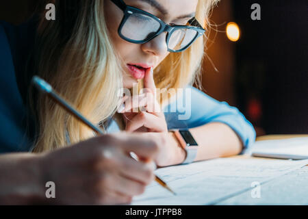 Bionda e giovane studente caucasici in occhiali di pensare e di scrivere qualcosa con la matita, lo studio fino a tardi Foto Stock