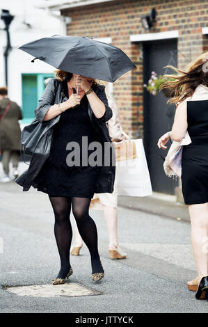 Brighton, Regno Unito. 10 Ago, 2017. Questa giovane donna ha problemi con il suo ombrello come lei si blocca sul suo cappello come essi arrivano vestita per l'occasione su un brillante ma breezy day a Brighton via gare Ladies Mobile giorno durante i tre giorni di Festival Maronthonbet di credito Racing: Simon Dack/Alamy Live News Foto Stock