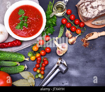 Il Gazpacho spagnolo zuppa fredda in una circolare bianca con piastra in ceramica su uno sfondo nero, accanto a verdure fresche per la cottura e il pane di segale su una lavagna Foto Stock