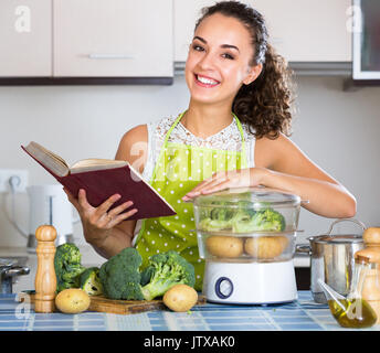 Allegro ragazza con libro culinaria e la cottura a vapore di verdure per cena salutare Foto Stock