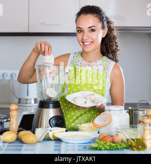 Ritratto di giovane sorridente donna fare crocchette di purè di patate con blender a casa Foto Stock