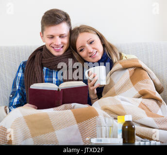 Congelati giovane seduto sul divano sotto una coperta con libro Foto Stock