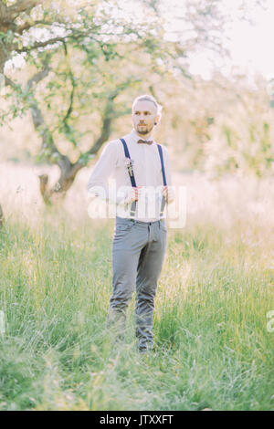 Lo sposo in pantaloni grigi e camicia bianca tenendo la sua black autoreggenti nel verde campo primaverile. Foto Stock