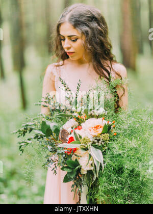 Close-up ritratto di donna con lunghi capelli ricci in matrimonio abito rosa con il bouquet di fiori colorati e guardando l'erba nella foresta. Foto Stock