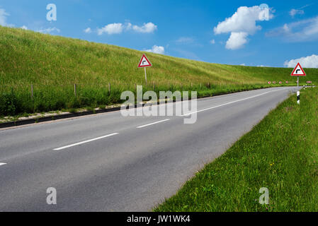 Segnale di avvertimento per indicare sharp curva a destra su entrambi i lati della strada vuota dietro una diga coperte di lussureggiante verde erba sotto il cielo di estate blu Foto Stock