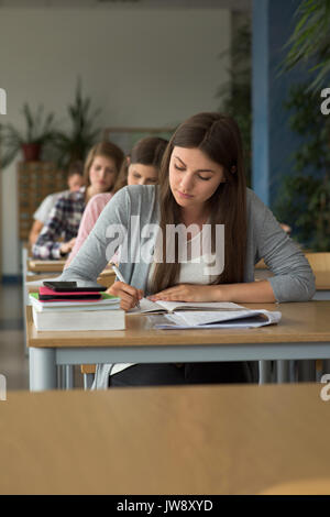 Studenti universitari la scrittura sul libro mentre è seduto alla scrivania durante l'esame in aula Foto Stock
