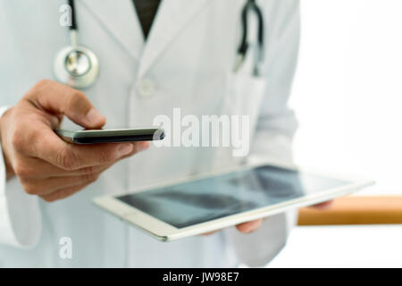 Primo piano di un giovane medico caucasica uomo che indossa un mantello bianco utilizzando uno smartphone mentre si sta verificando una radiografia del torace in un computer tablet Foto Stock