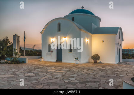 Una piccola chiesa bianca sulla spiaggia a Paphos, Cipro durante l'alba. Il sole sorge sopra l'orizzonte e illumina la chiesa. Foto Stock