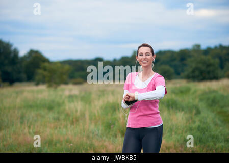 Un sano, donna sorridente nel montare l'usura con smart phone, guardare e auricolari, esercitare all'aperto in natura. Foto Stock