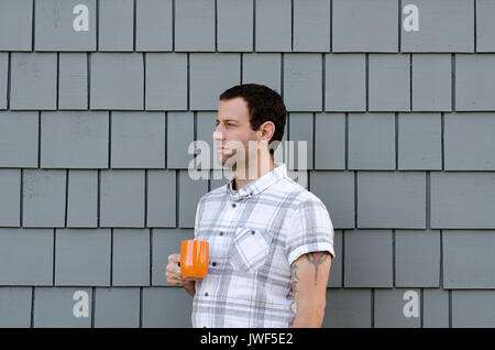Profilo laterale di un attraente uomo che indossa un plaid bianco shirt tenendo un arancio tazza da caffè contro uno sfondo grigio. Foto Stock