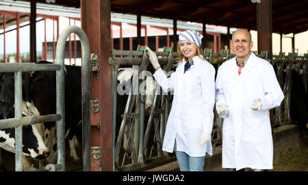Allegro dei salariati agricoli in piedi vicino alla mungitura allevamento nel granaio e sorridente Foto Stock