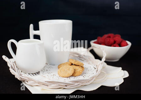 Tazza di caffè, biscotti una brocca del latte su un vassoio di paglia Una ciotola con lamponi. Sfondo nero Foto Stock