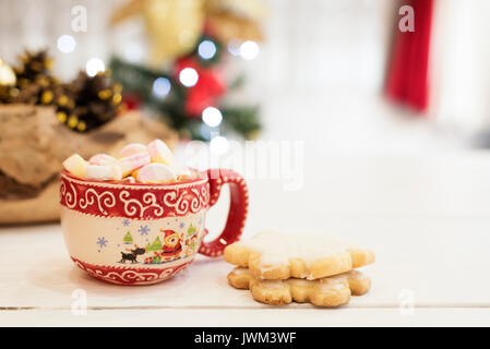 La cioccolata calda con marshmallow caramelle. Biscotti di Natale in forma di fiocchi di neve, golden coni e albero di natale luci. Bianco sullo sfondo di legno Foto Stock