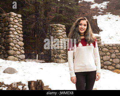 Una sorridente ragazza teen in bianco e rosso maglione da un cancello di ferro con pilastri di pietra in inverno Foto Stock