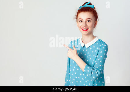 Guardate questo! Felice allegro giovani caucasici redhead ragazza con i capelli bun puntando il suo dito indice di distanza, indicando copia su uno spazio vuoto bianco parete per Foto Stock