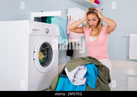 Giovane donna infelice guardando il mucchio di vestiti sporchi vicino alla macchina di lavaggio in lavanderia Foto Stock