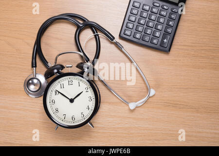 Elevato angolo di visione di uno stetoscopio acustico tra sveglia con la calcolatrice contro lo sfondo di legno Foto Stock