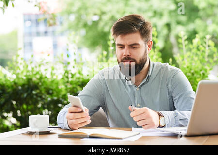 Ritratto di giovane imprenditore barbuto utilizza lo smartphone mentre si lavora al cafe' all'aperto Foto Stock