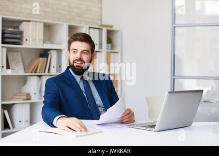Ritratto di successo imprenditore barbuto sorridente felicemente alla ricerca di distanza mentre si lavora alla scrivania in ufficio moderno Foto Stock