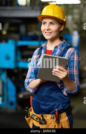 Ritratto di donna operaio di fabbrica indossando hardhat utilizzando tavoletta digitale e sorridente, guardando lontano Foto Stock