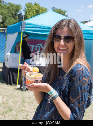 Giovane donna con dritto marrone capelli e occhiali da sole sorridente e mangiare di colore arancio gelato Austin Gelato Festival. Tenda esterna dietro. Foto Stock