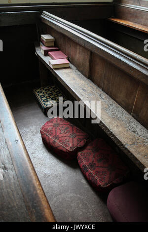 Vecchia chiesa pew illuminato dalla luce della finestra corre diagonalmente attraverso il telaio: hymnals impilati su una estremità e il rosso e il verde inginocchiatoi modellato sotto. Foto Stock