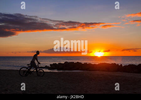 Un giovane mam spinge la sua bicicletta lungo la spiaggia come il sole tramonta a Port St Charles, Barbados Foto Stock