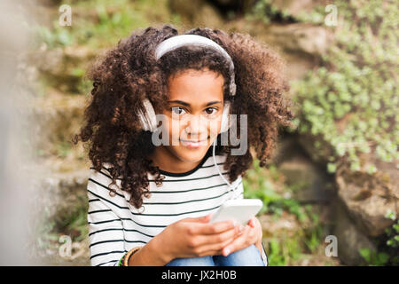 Bella americano africano ragazza con i capelli ricci all'aperto, seduti su scale, tenendo smart phone, indossando le cuffie e ascoltare della musica. Foto Stock