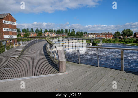 Un ponte pedonale che attraversa un fiume con uno stramazzo a th lato. Foto Stock