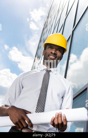 Basso angolo vista di architetto professionista in hard hat holding blueprint al di fuori di un edificio moderno Foto Stock