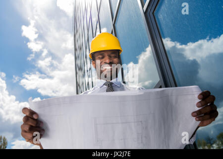 Basso angolo vista di architetto professionista in hard hat holding blueprint al di fuori di un edificio moderno Foto Stock