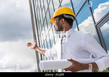 Basso angolo vista di architetto professionista in hard hat holding blueprint al di fuori di un edificio moderno Foto Stock