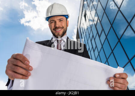 Basso angolo di vista sorridente architetto barbuto holding blueprint e guardando la fotocamera Foto Stock