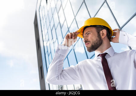 Vista laterale del fiducioso architetto professionista in hard hat contro la costruzione di Foto Stock