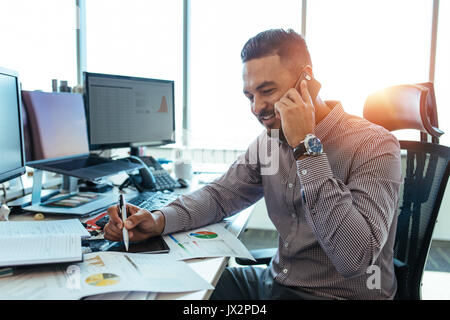 uomo d'affari che parla al telefono cellulare e guarda in basso con un sorriso che indica i documenti aziendali. Uomo seduto alla scrivania del suo ufficio a lavorare su grafici e caratteri Foto Stock